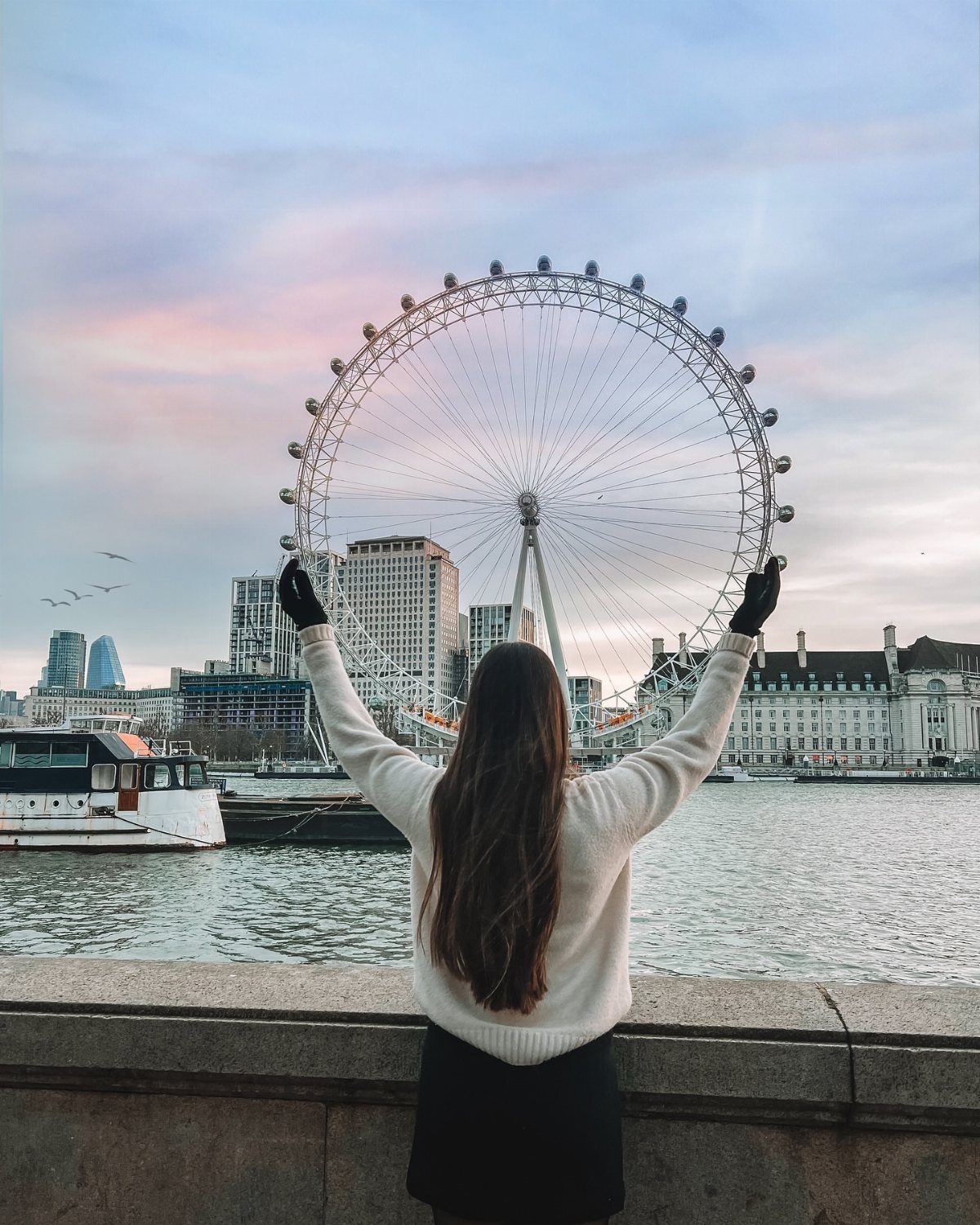 Woman with arms raised facing a large observation wheel across a city river.