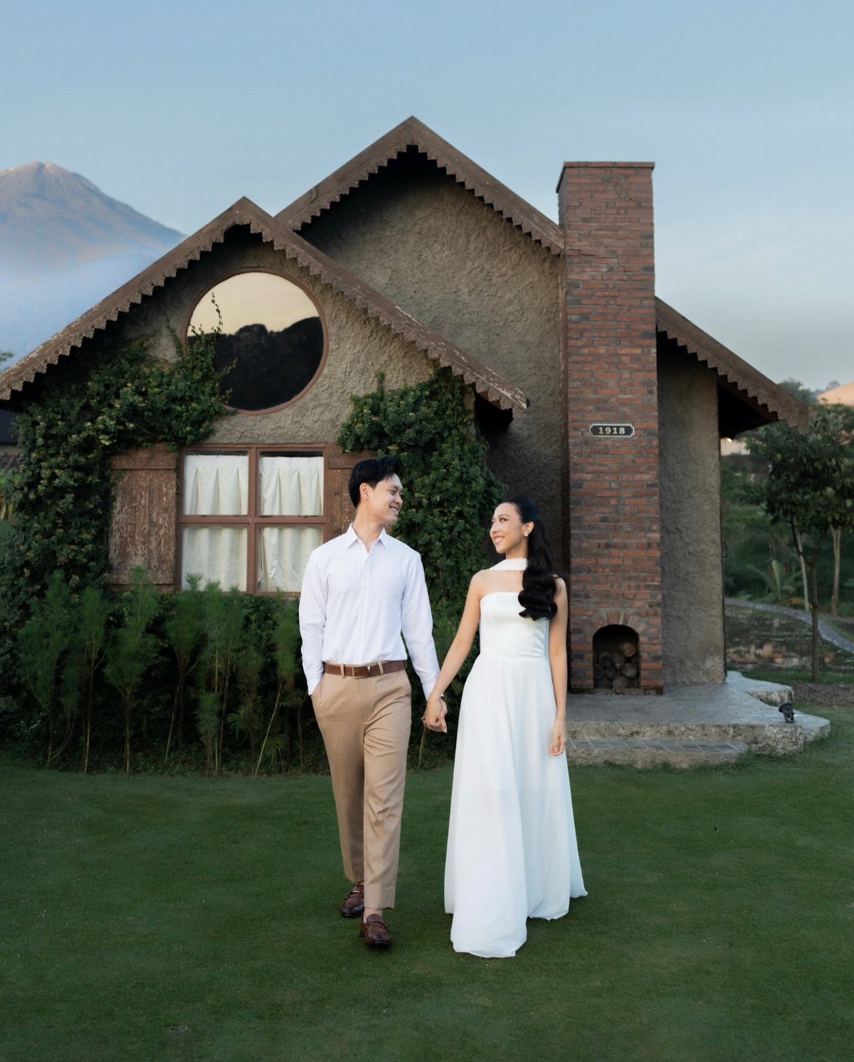 Couple in white attire walking hand-in-hand on a lawn before a rustic house.