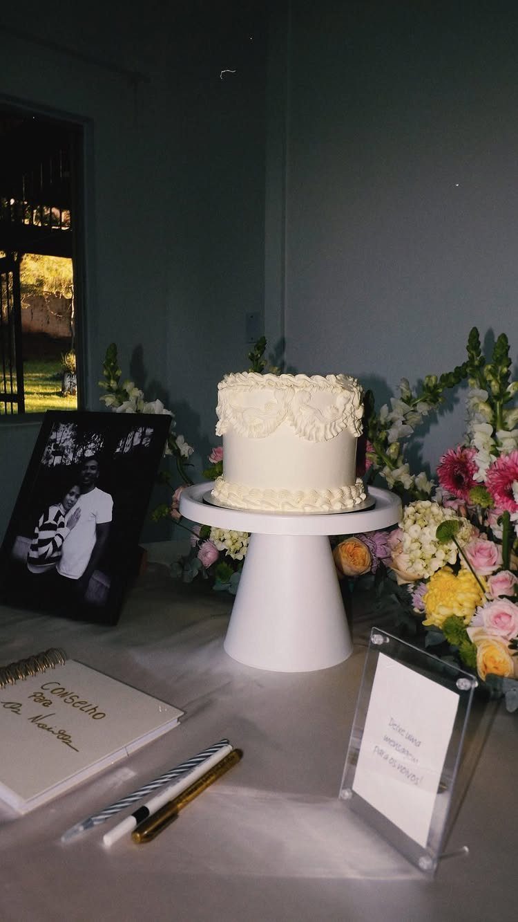 White frosted cake on a pedestal stand next to flowers and a guest book.