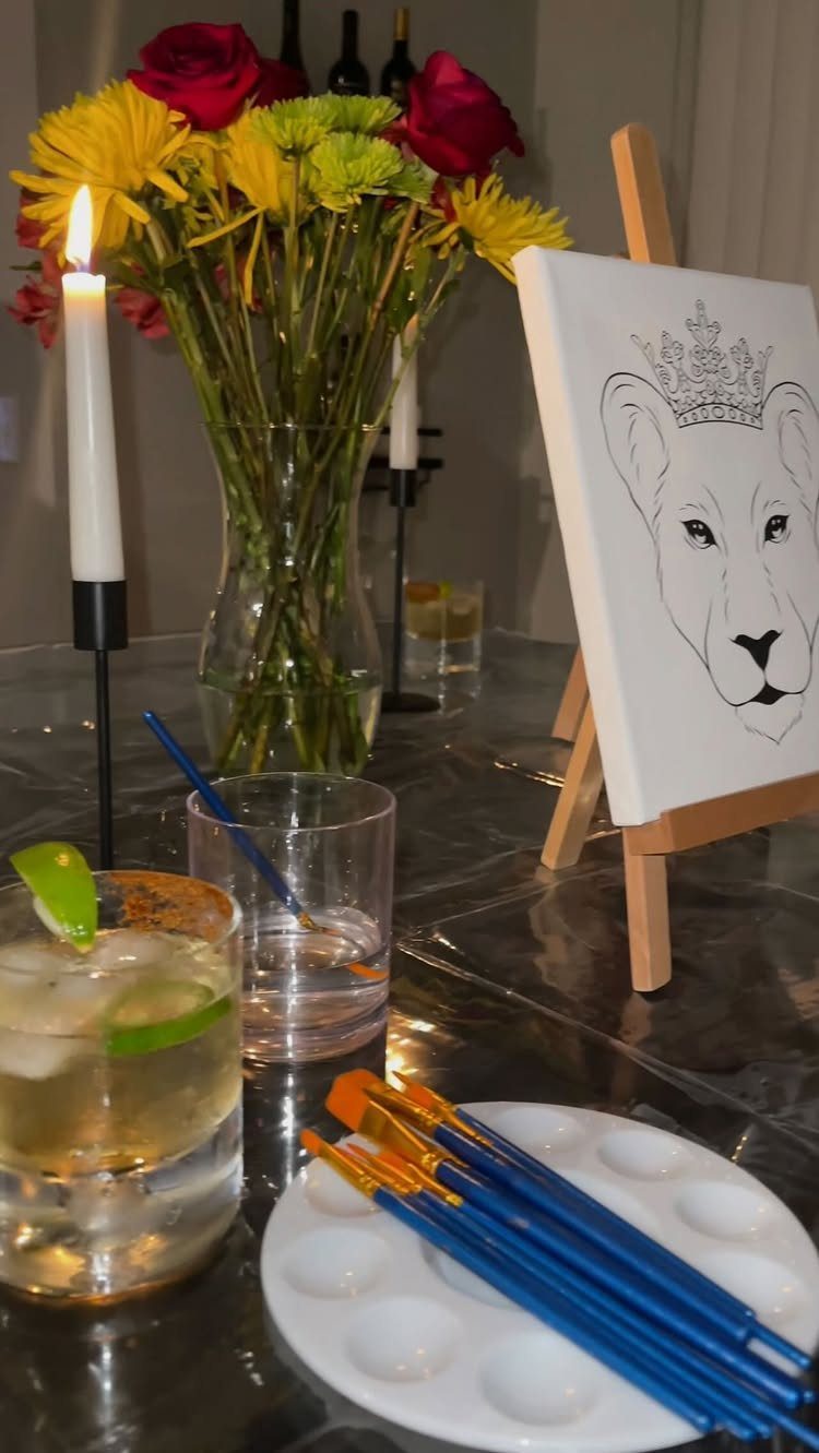 Canvas with lion drawing on an easel next to paintbrushes, palette, and cocktails.