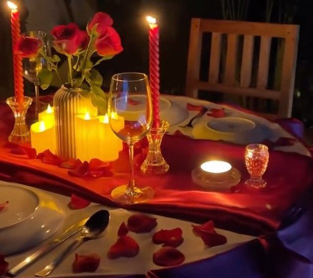 Romantic table setting with red roses, lit candles, and rose petals on red cloth.