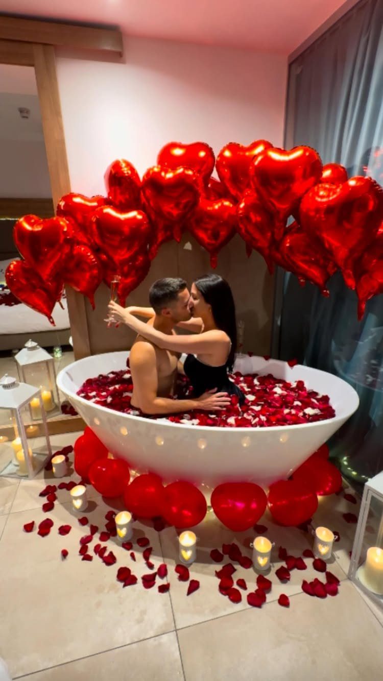 Couple kissing in a bathtub filled with red rose petals and heart balloons.