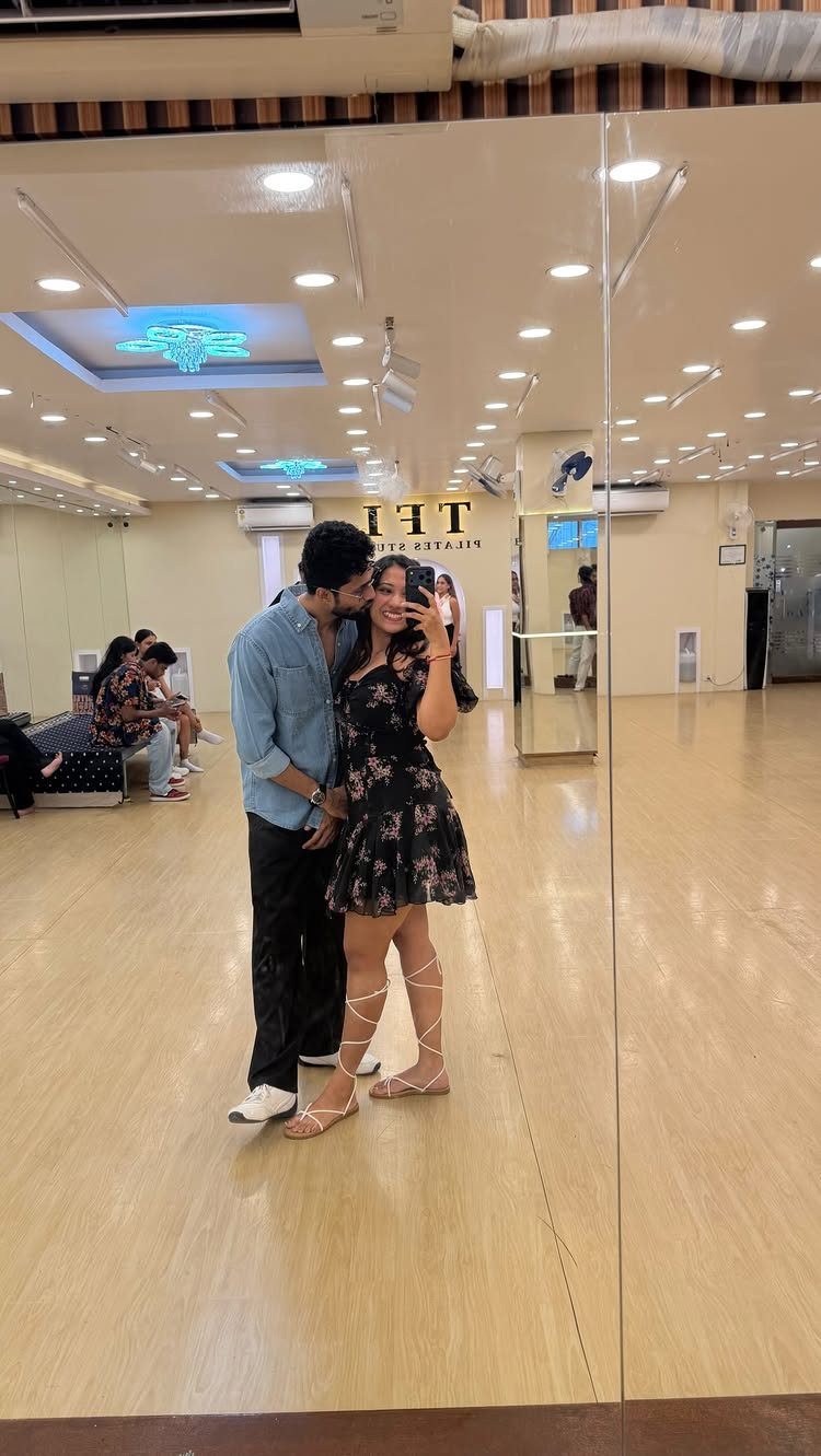 Couple posing for a mirror selfie inside a bright dance studio with wooden floors.