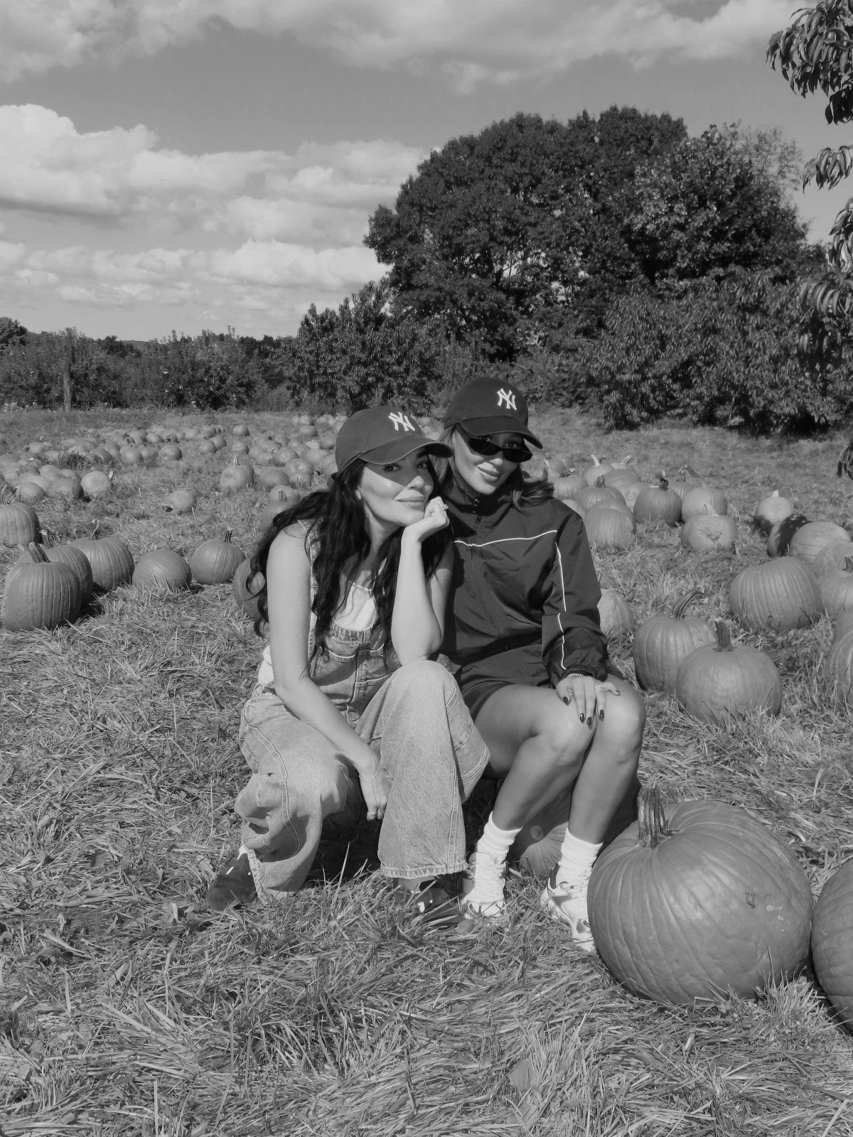Two women in baseball caps crouching in a pumpkin patch with many pumpkins.