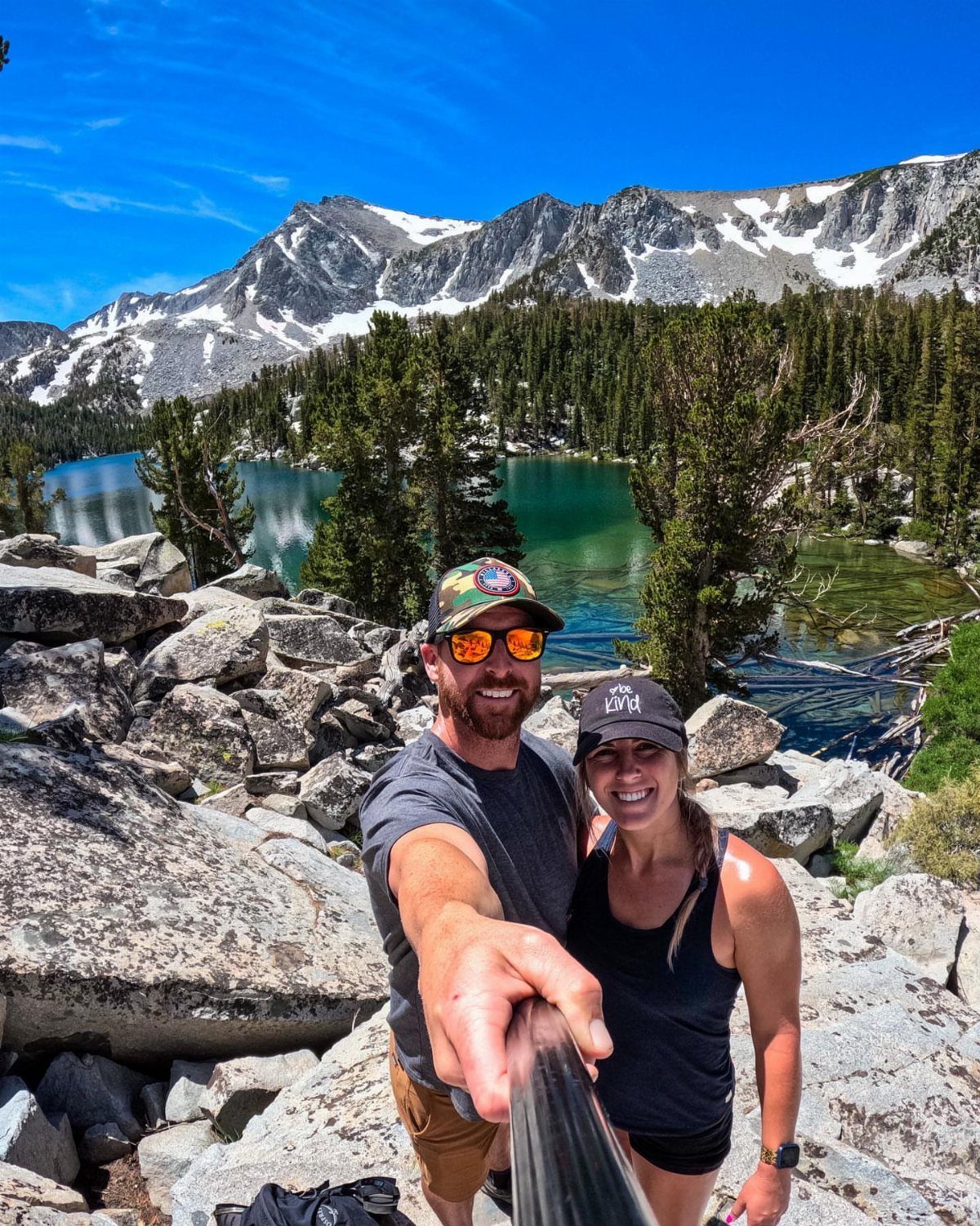 Smiling couple taking a selfie in front of a mountain lake and peaks.