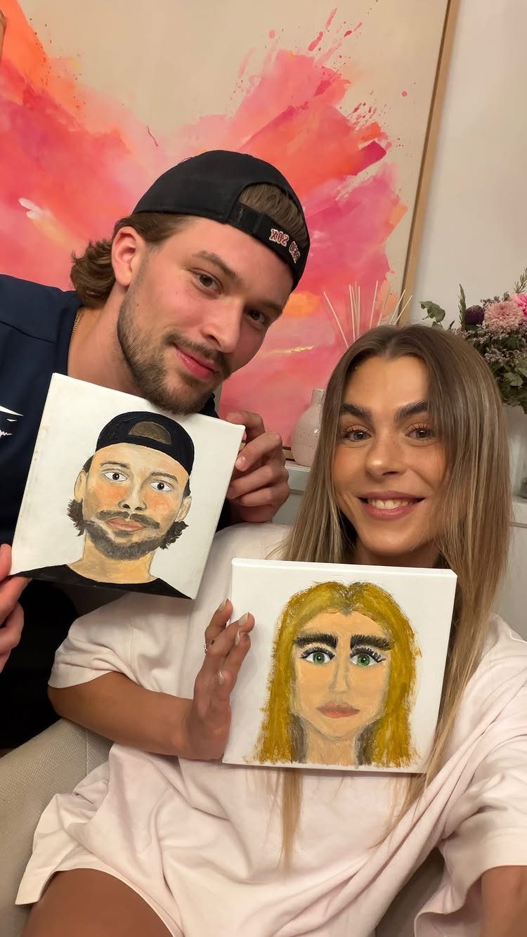 Couple holding hand-painted portraits of each other while smiling for the camera indoors.