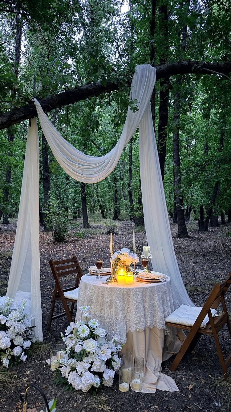 Romantic dinner table with white lace and candles set under a forest tree canopy