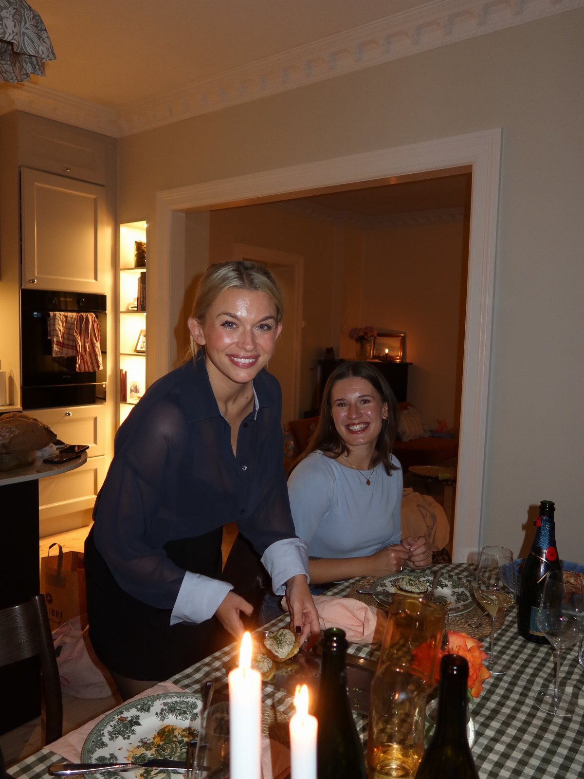 Two women smiling behind a dinner table with candles and wine in a home.