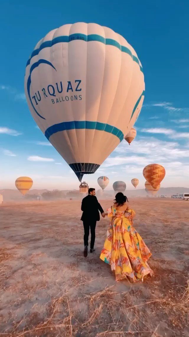 Couple holding hands in a field with several hot air balloons in the sky