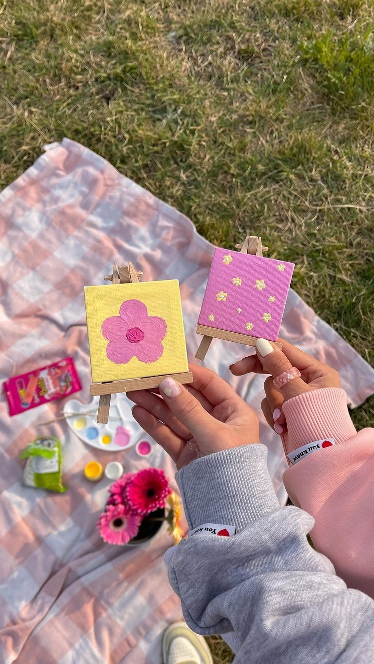Two people painting small flower canvases on a picnic blanket in a park.