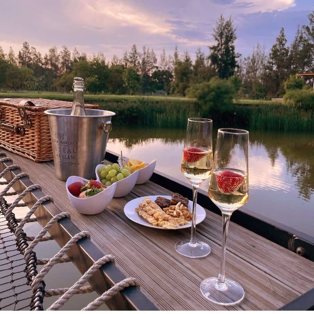 Picnic setup with champagne, fruit, and snacks on a deck overlooking a lake.