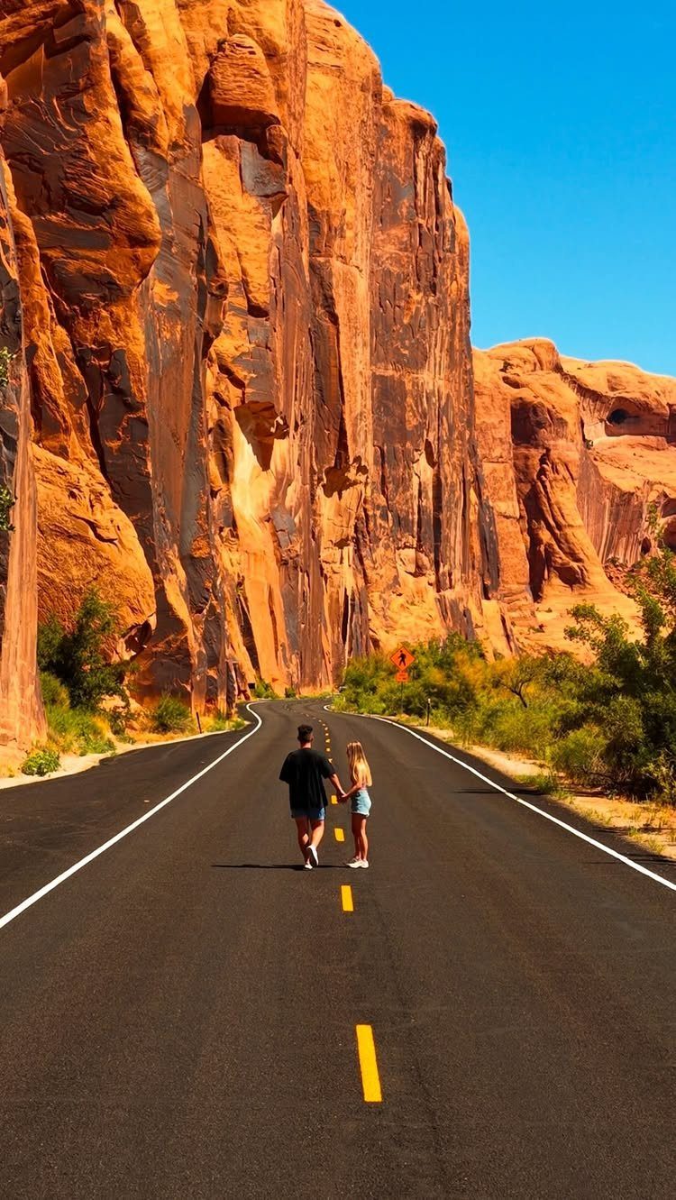 Couple walking hand-in-hand on a road surrounded by massive red rock cliffs.