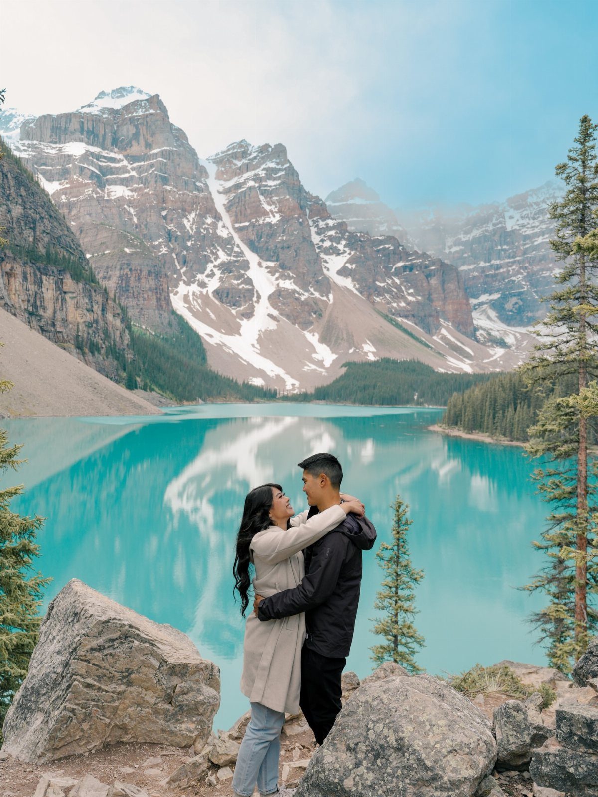 Couple embracing on a rocky cliff overlooking a turquoise lake and snowy mountains.