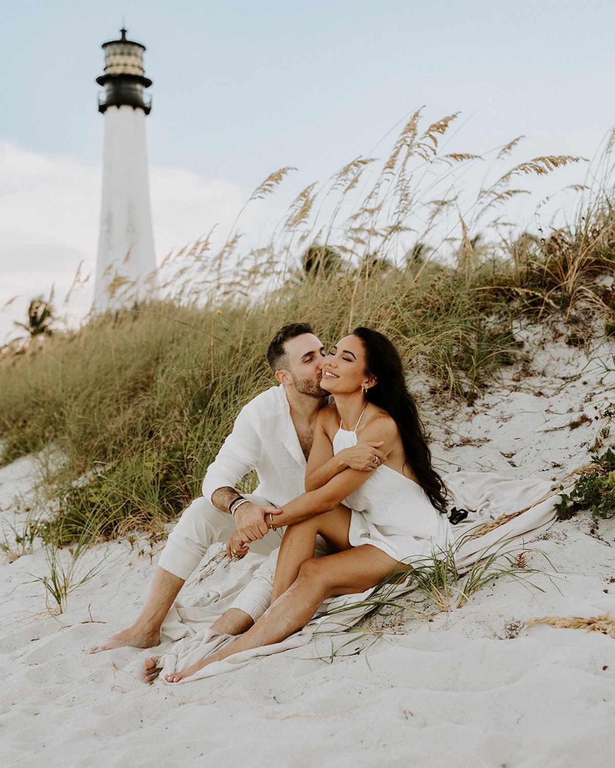 Couple in white clothing cuddling on a white sand beach with a lighthouse.