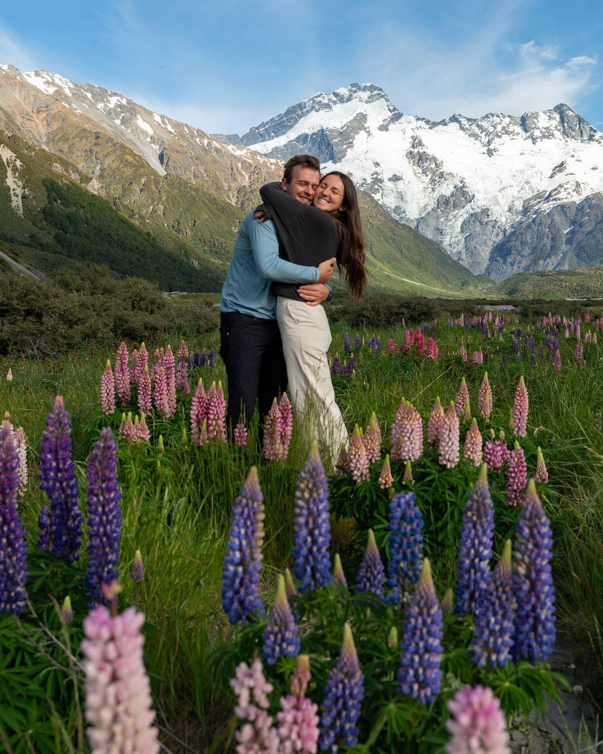 Couple embracing in a field of purple lupines with snow-capped mountains behind them.
