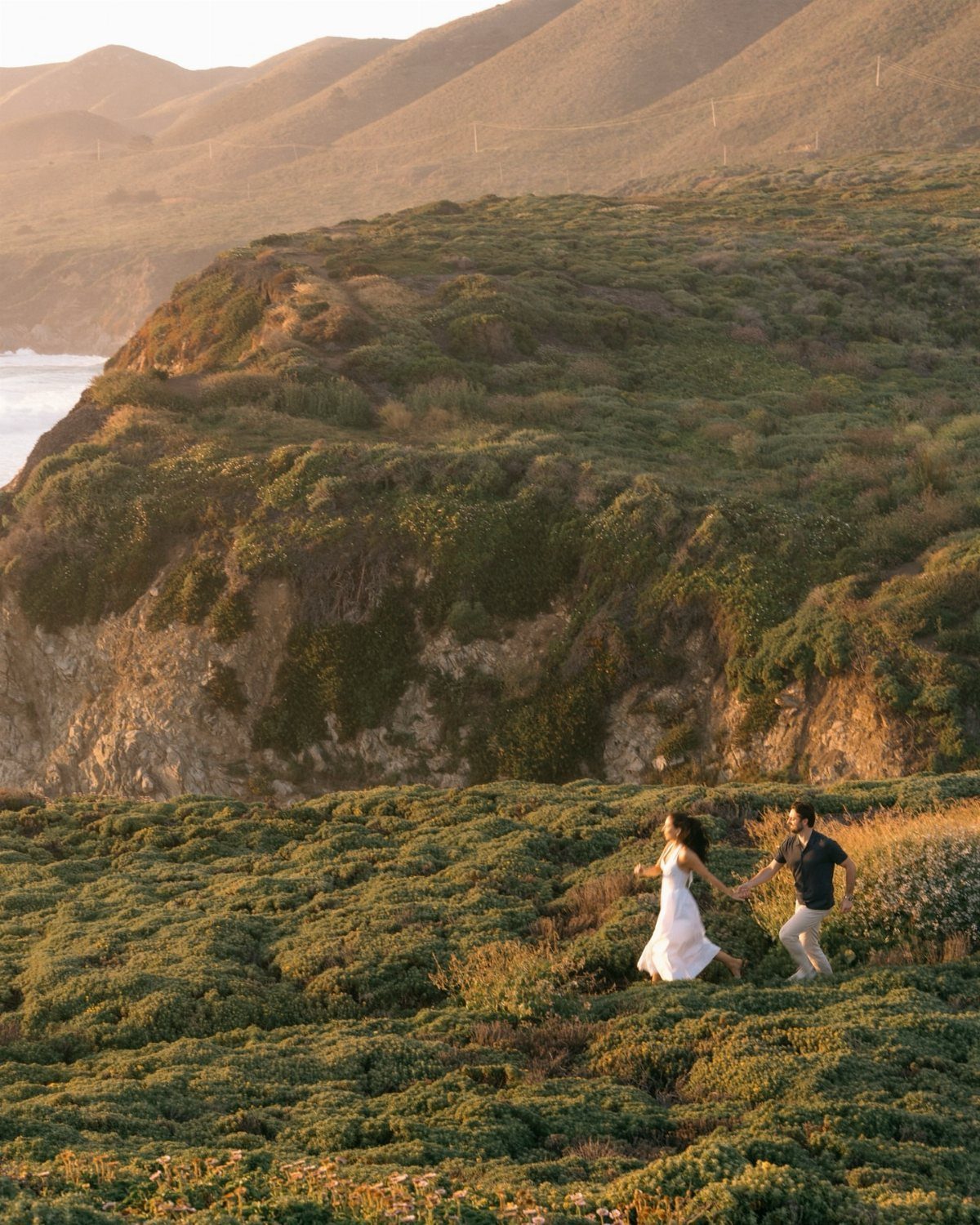 Couple holding hands walking through green coastal hills overlooking the ocean