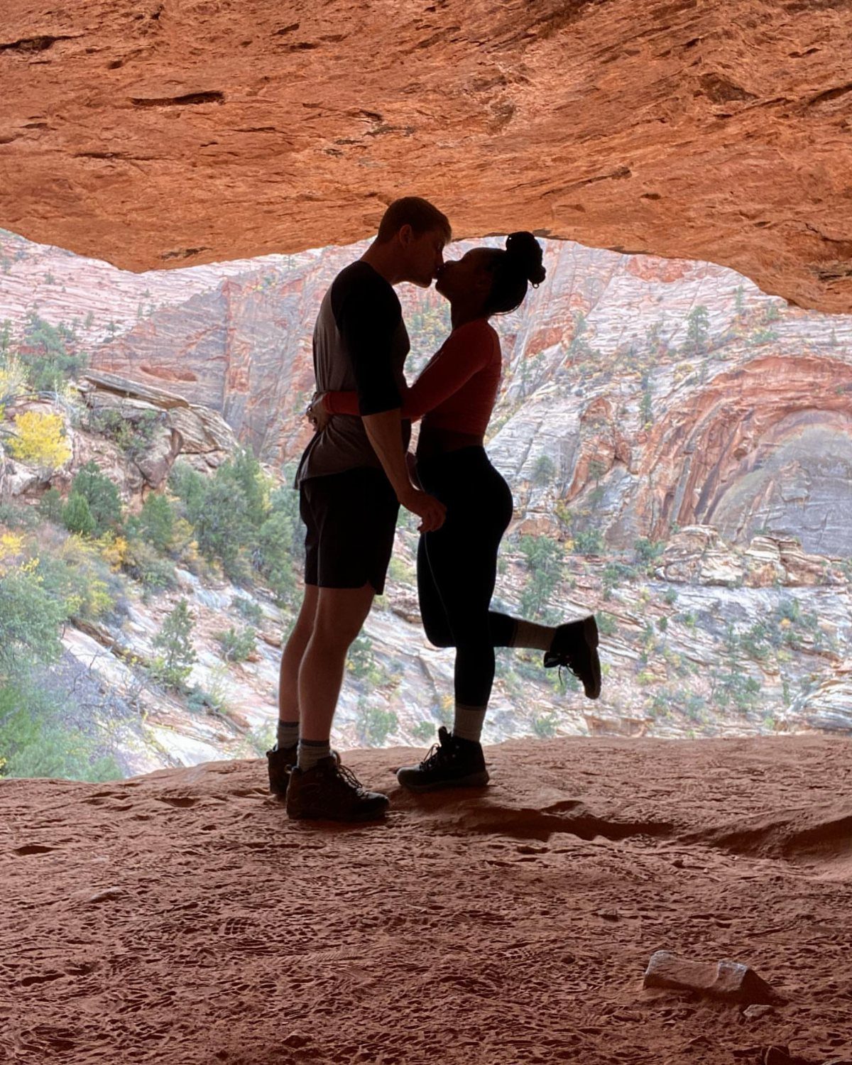 Couple kissing in silhouette under a red rock arch overlooking a canyon landscape.