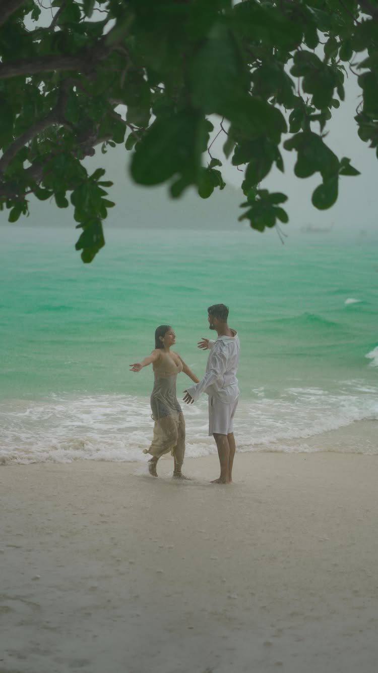 Couple standing on a white sand beach with turquoise water and green foliage.