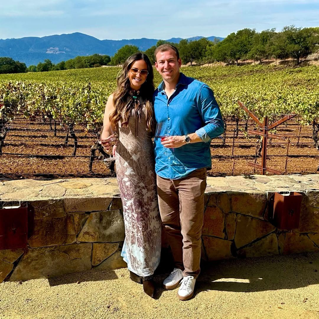 Couple smiling in front of a vineyard with rolling hills and mountains.