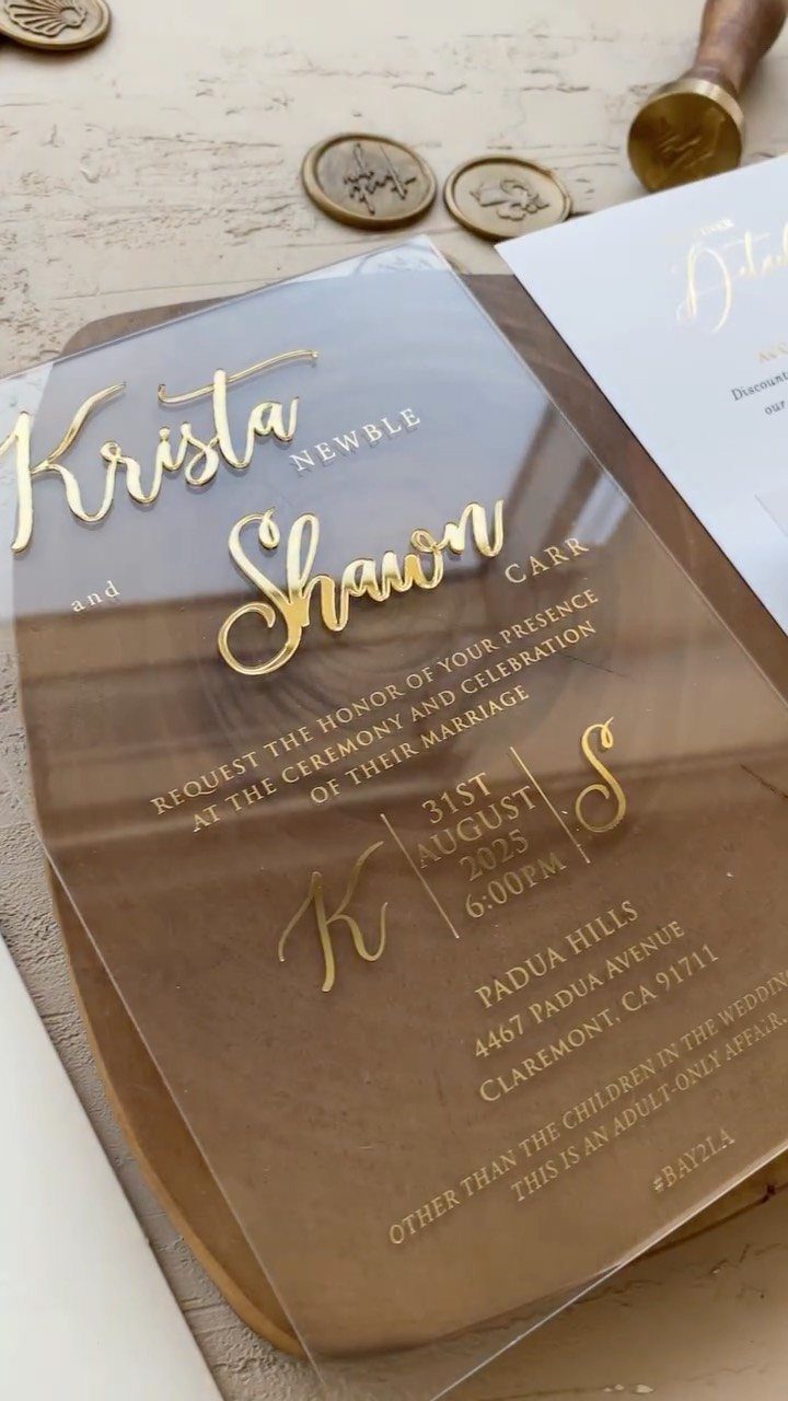 Clear acrylic wedding invitation with gold calligraphy and gold wax seals nearby.