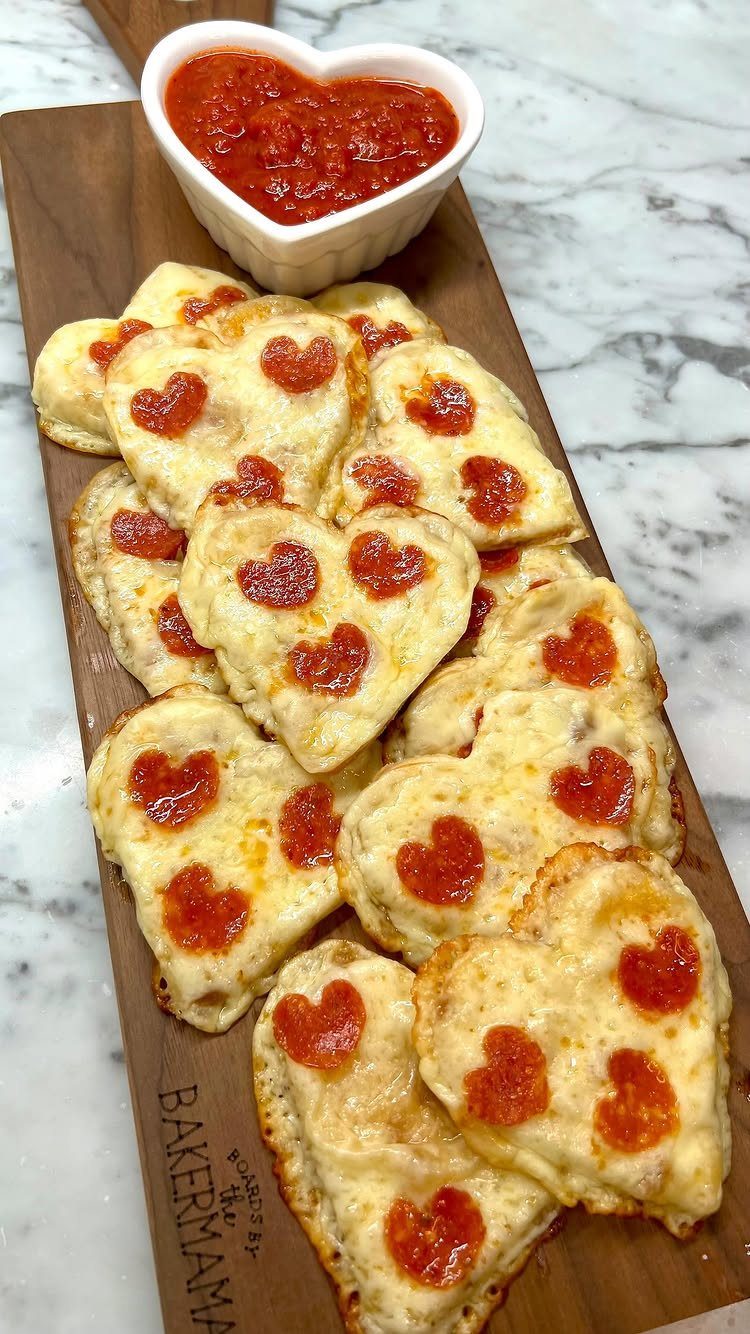 Heart-shaped mini pizzas with heart pepperoni and marinara sauce on a wooden board.