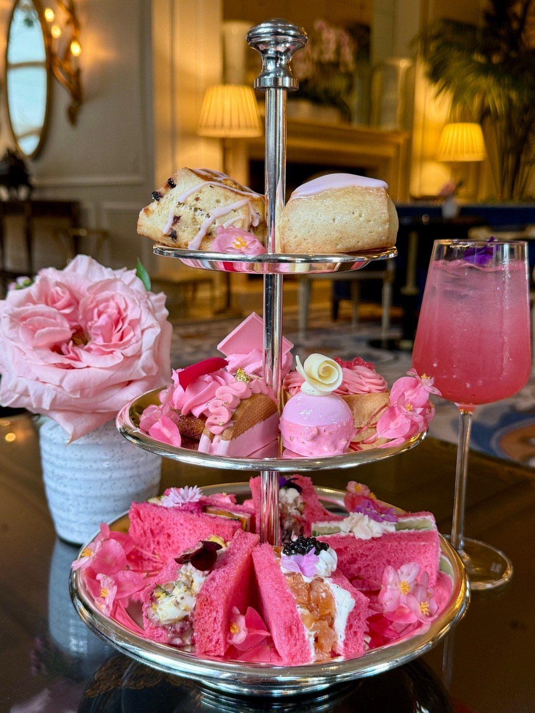 Three-tiered silver stand with pink cakes and sandwiches next to a pink cocktail.