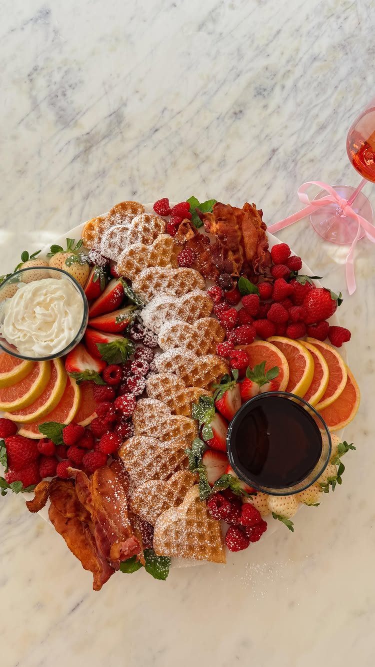 Heart-shaped brunch platter with waffles, fresh berries, oranges, and bacon on marble.