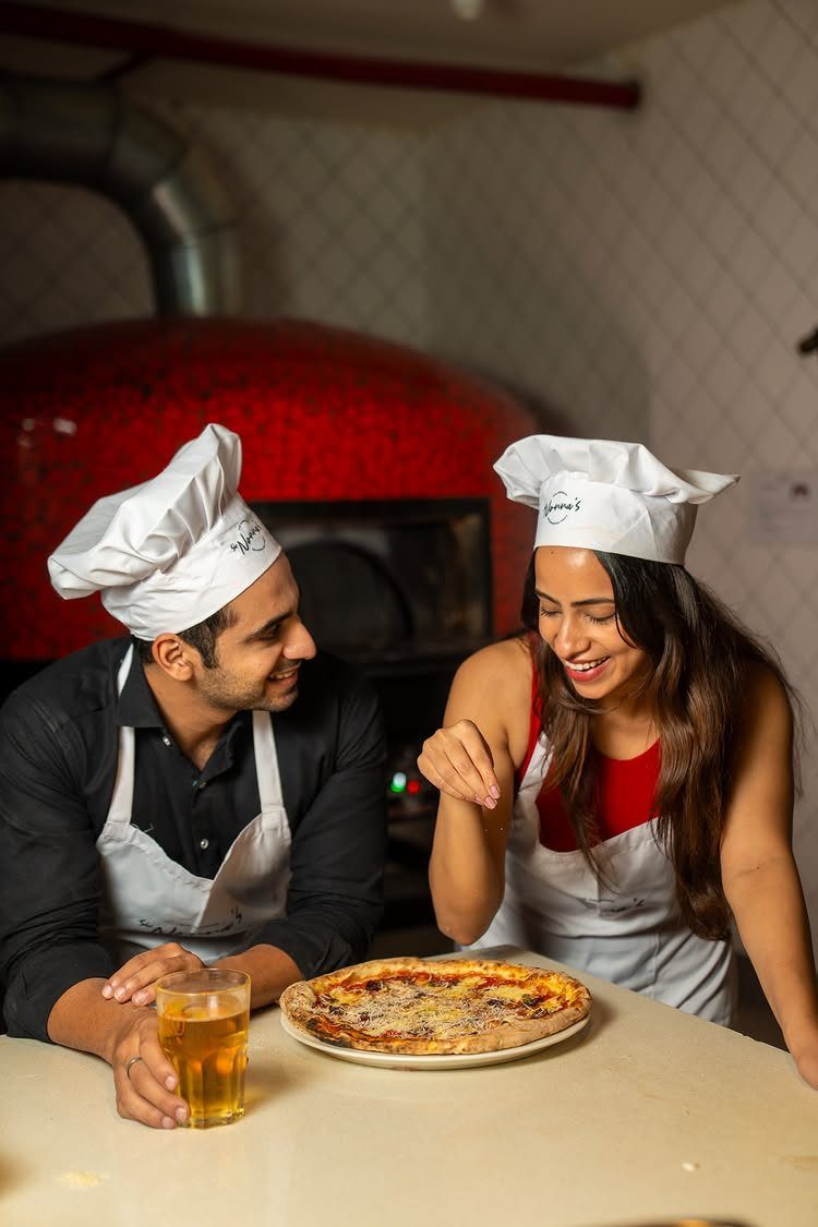 Couple in chef hats and aprons smiling over a pizza in a kitchen