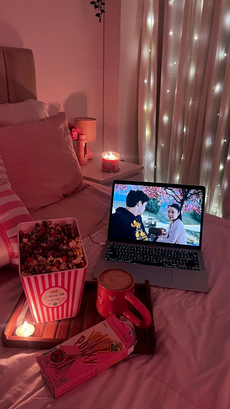 Cozy bedroom date setup with laptop, popcorn, and drinks on pink bedding