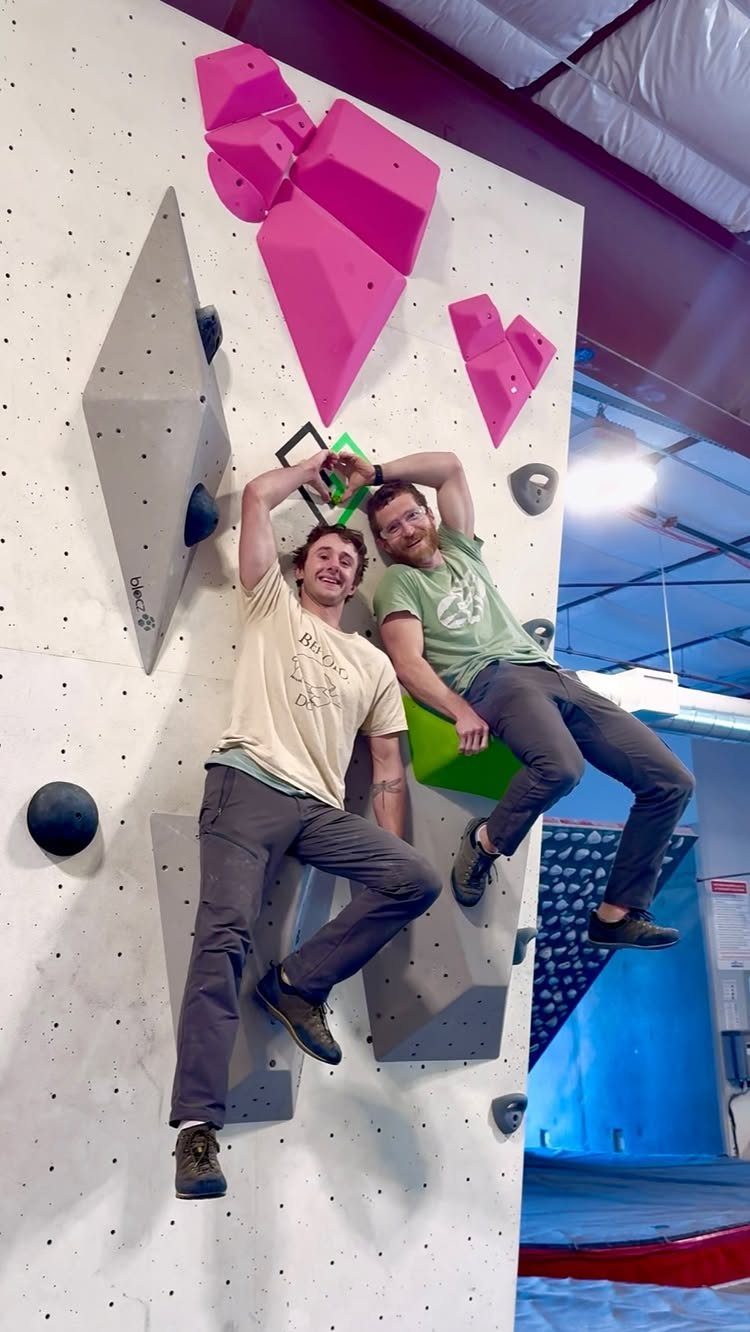 Two men smiling and holding hands while climbing an indoor bouldering wall.