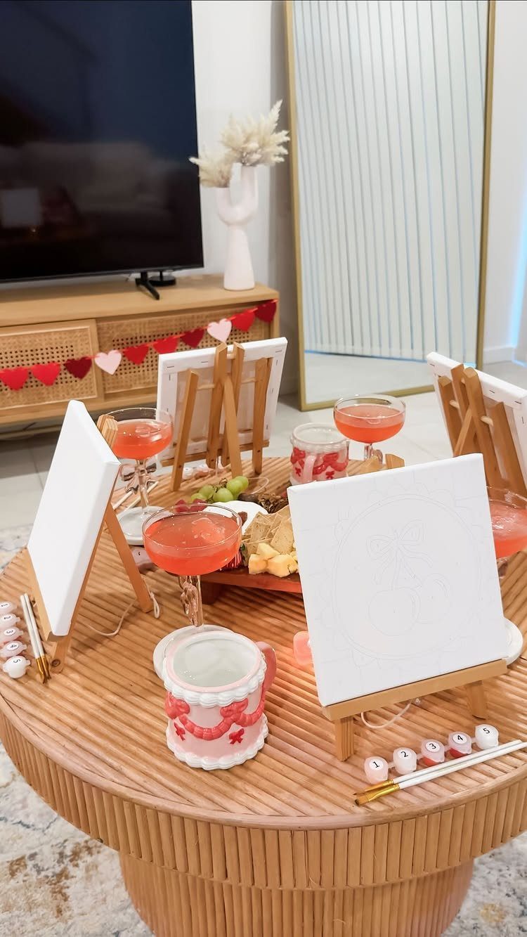 Painting date setup with canvases on easels, pink cocktails, and fruit on rattan table