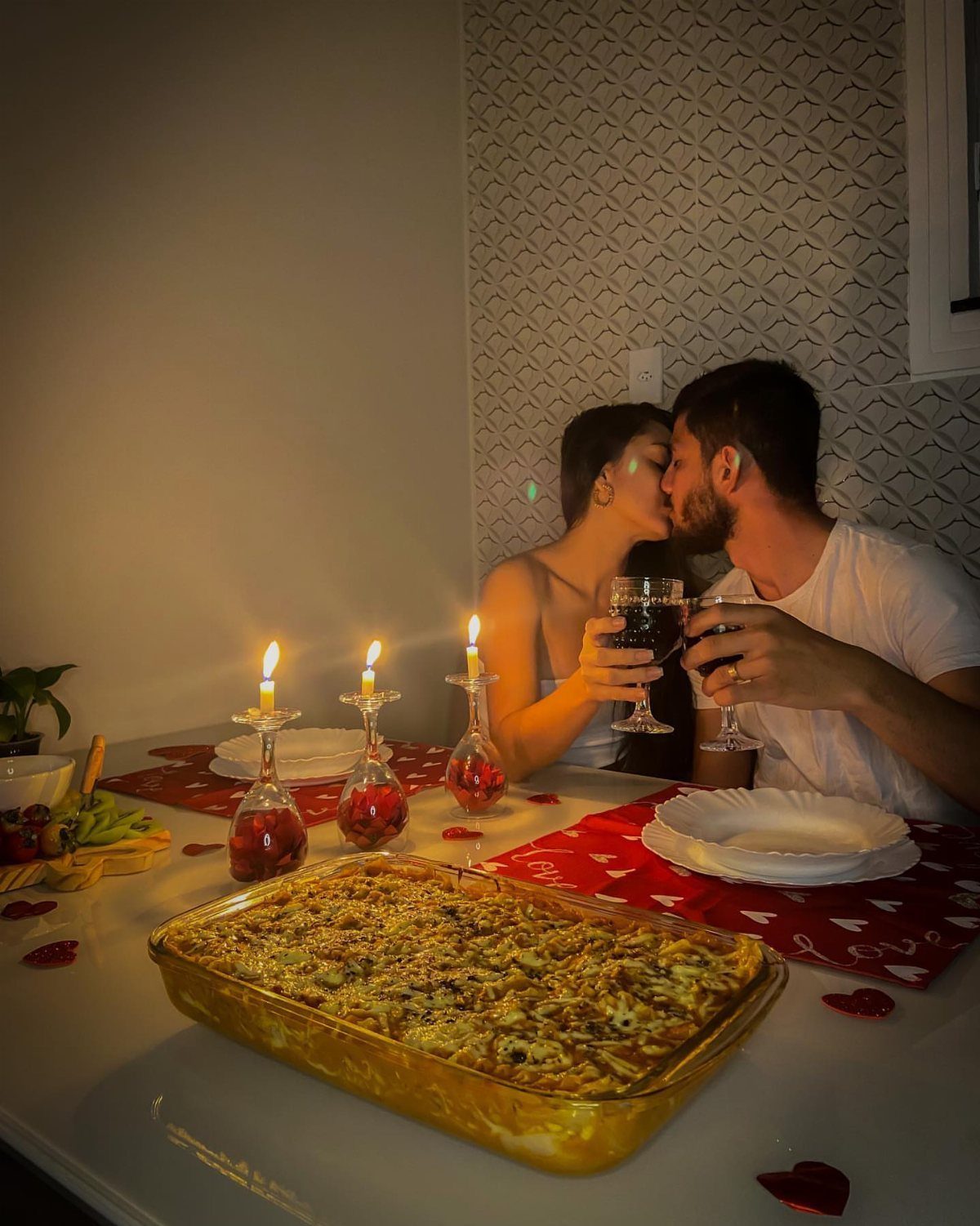 Couple kissing over red wine and a baked meal at a romantic dinner table.