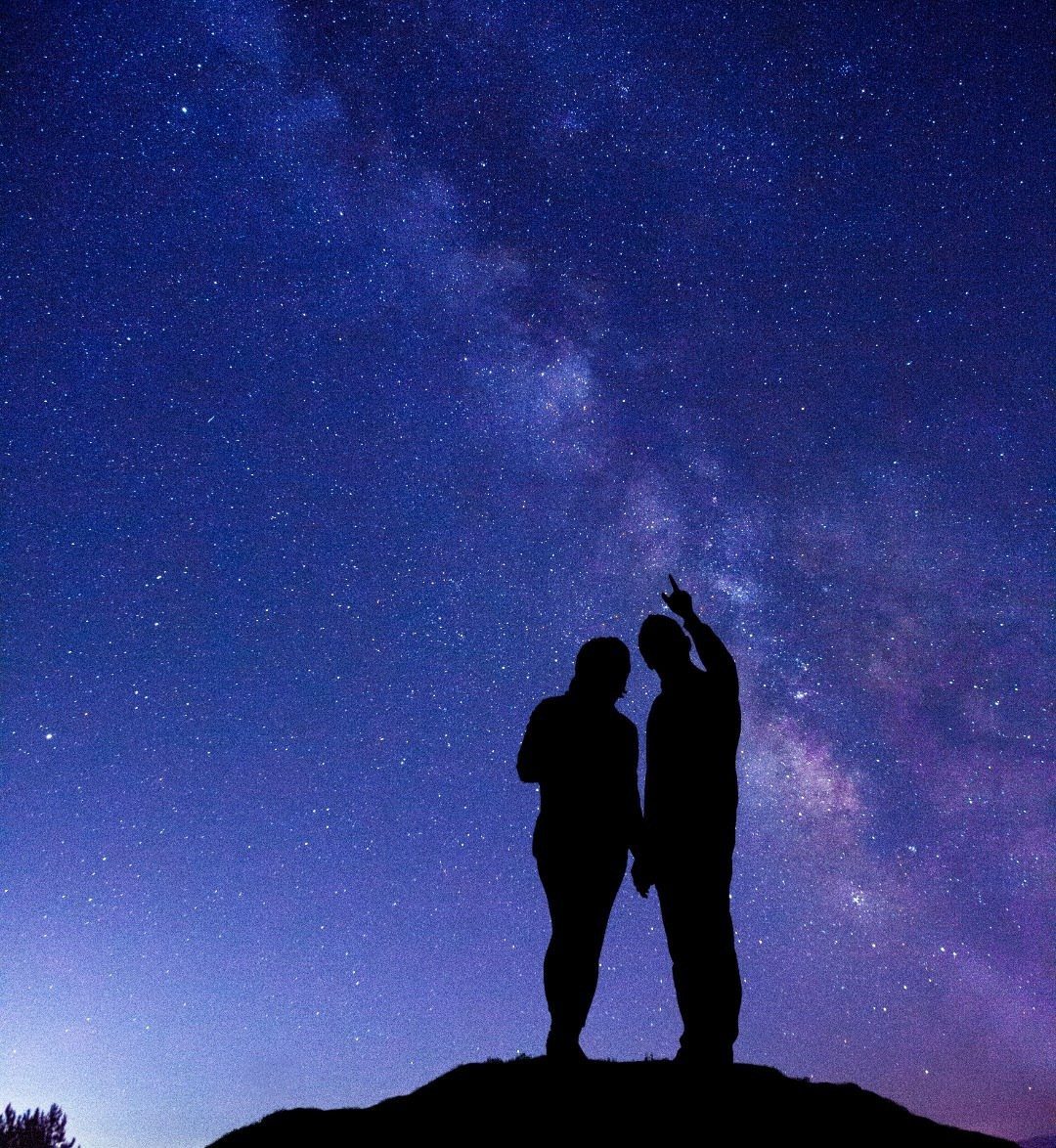 Silhouetted couple standing on a hill gazing at the starry Milky Way galaxy.