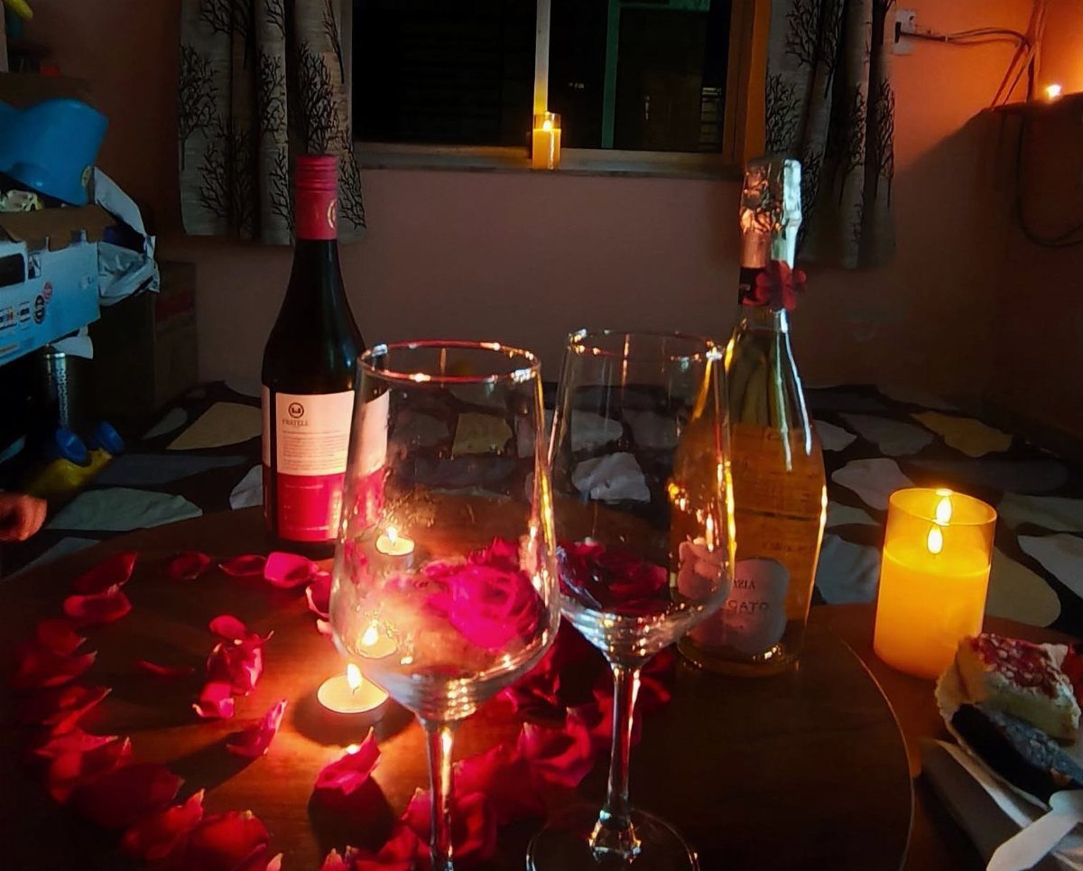 Wine bottles and glasses surrounded by red rose petals and glowing candles indoors.