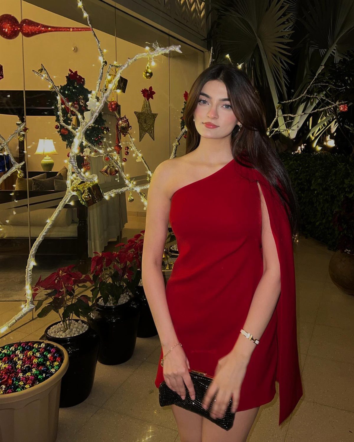 Woman in a one-shoulder red mini dress posing by a decorated holiday tree.