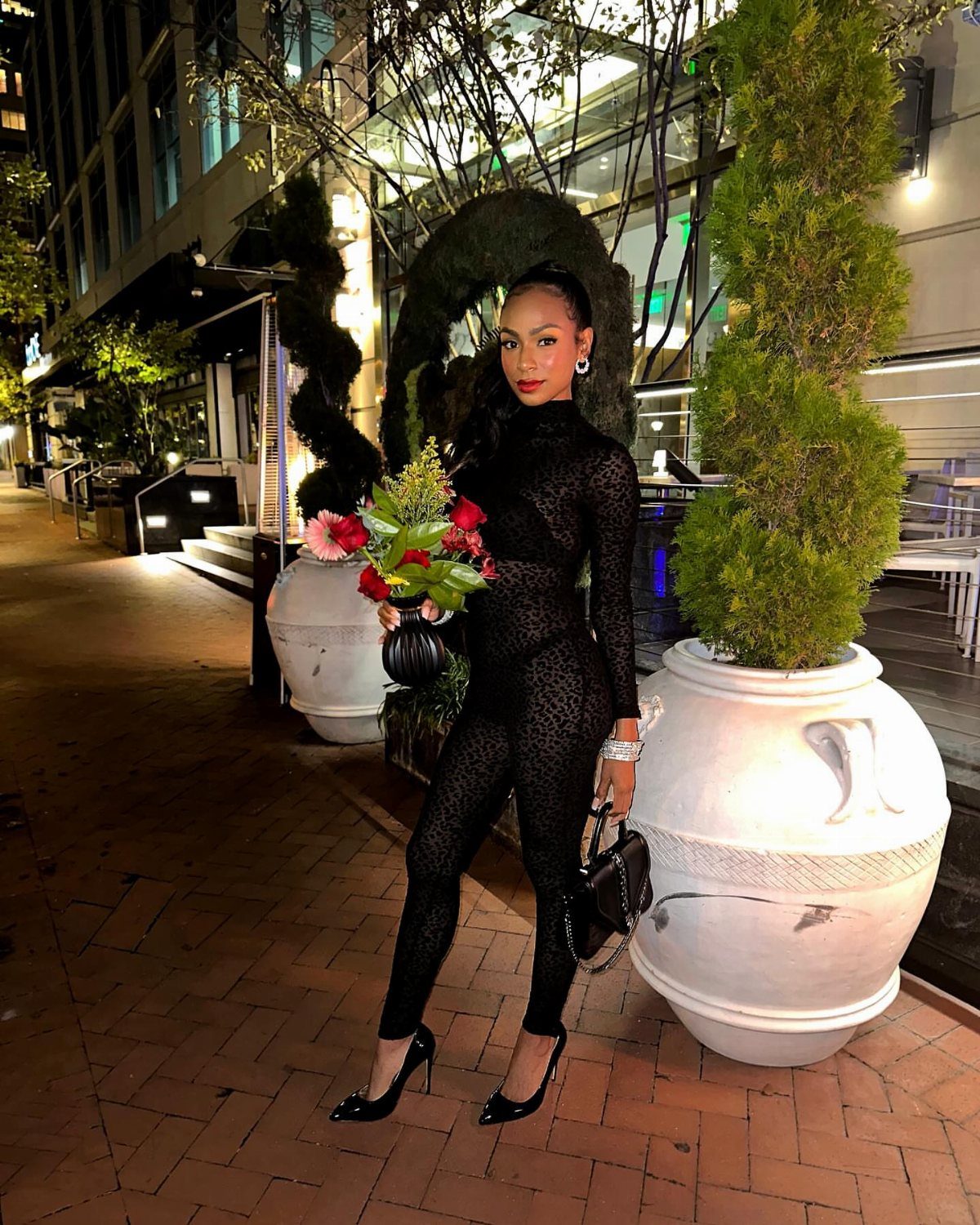 Woman in a black form-fitting jumpsuit holding red flowers on a city sidewalk