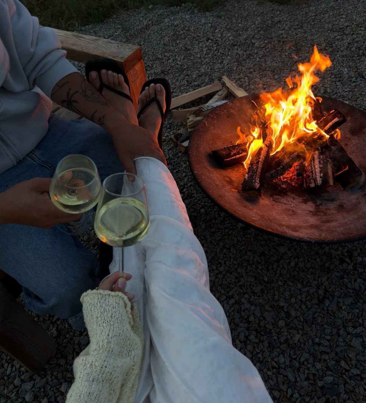 Couple holding glasses of white wine sitting beside a glowing outdoor bonfire pit.