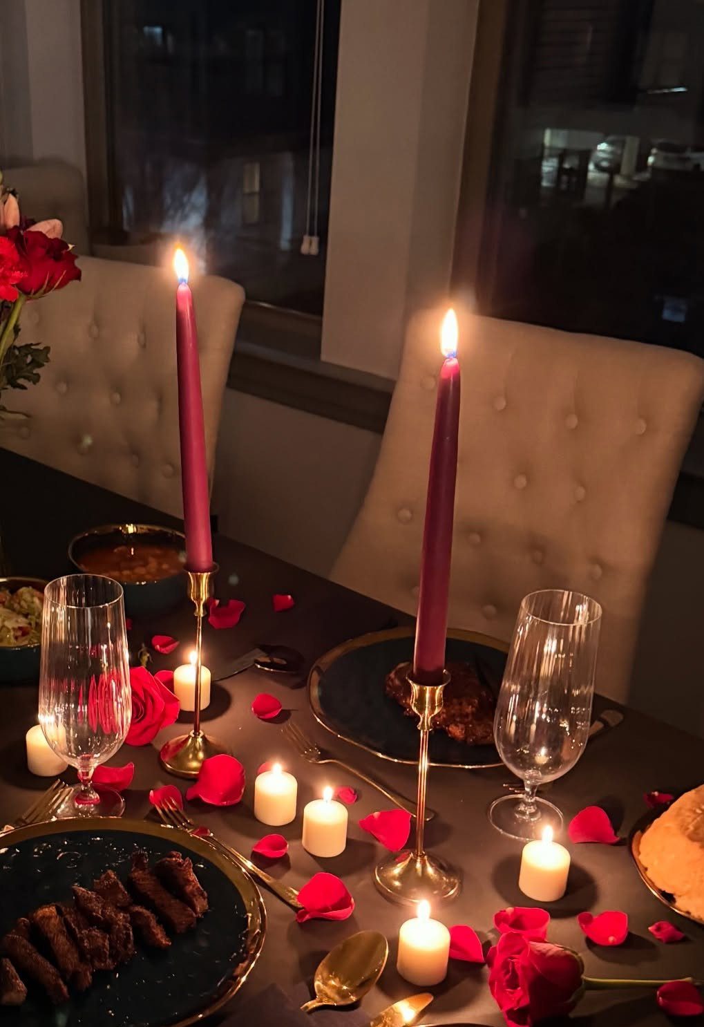 Romantic dinner table with pink taper candles, red rose petals, and gourmet food.
