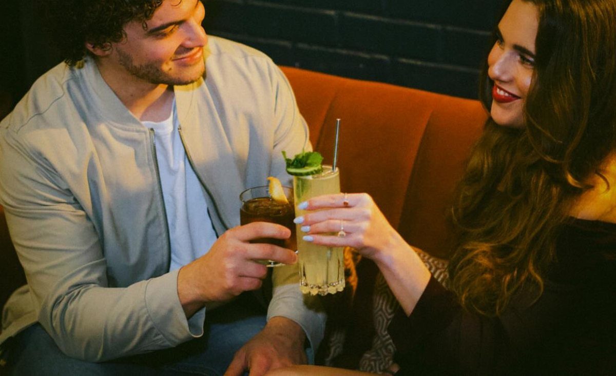 Couple clinking cocktail glasses while sitting on an orange couch in a bar.