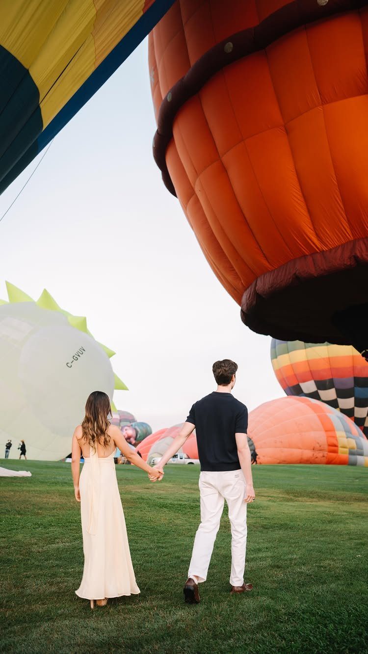 Couple holding hands walking through a field with colorful hot air balloons.