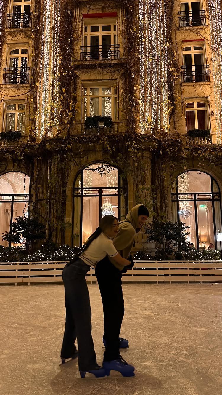 Couple ice skating together on an outdoor rink in front of a lit building.