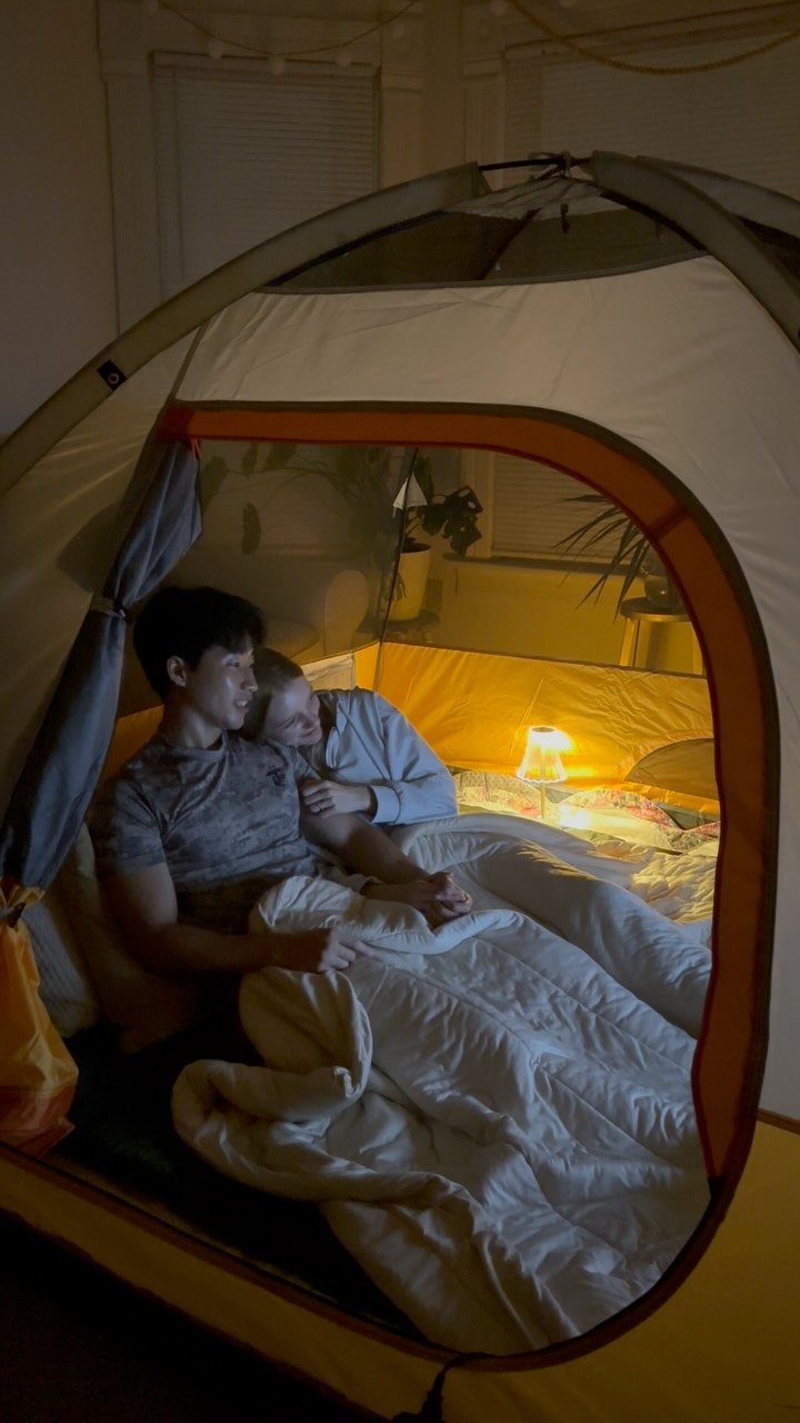 Couple cuddling under a duvet inside a camping tent set up indoors.