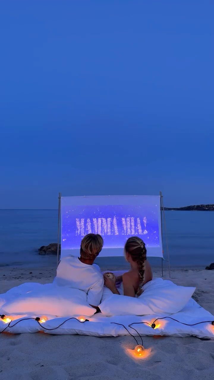 Couple watching a movie on a projector screen on a beach with pillows