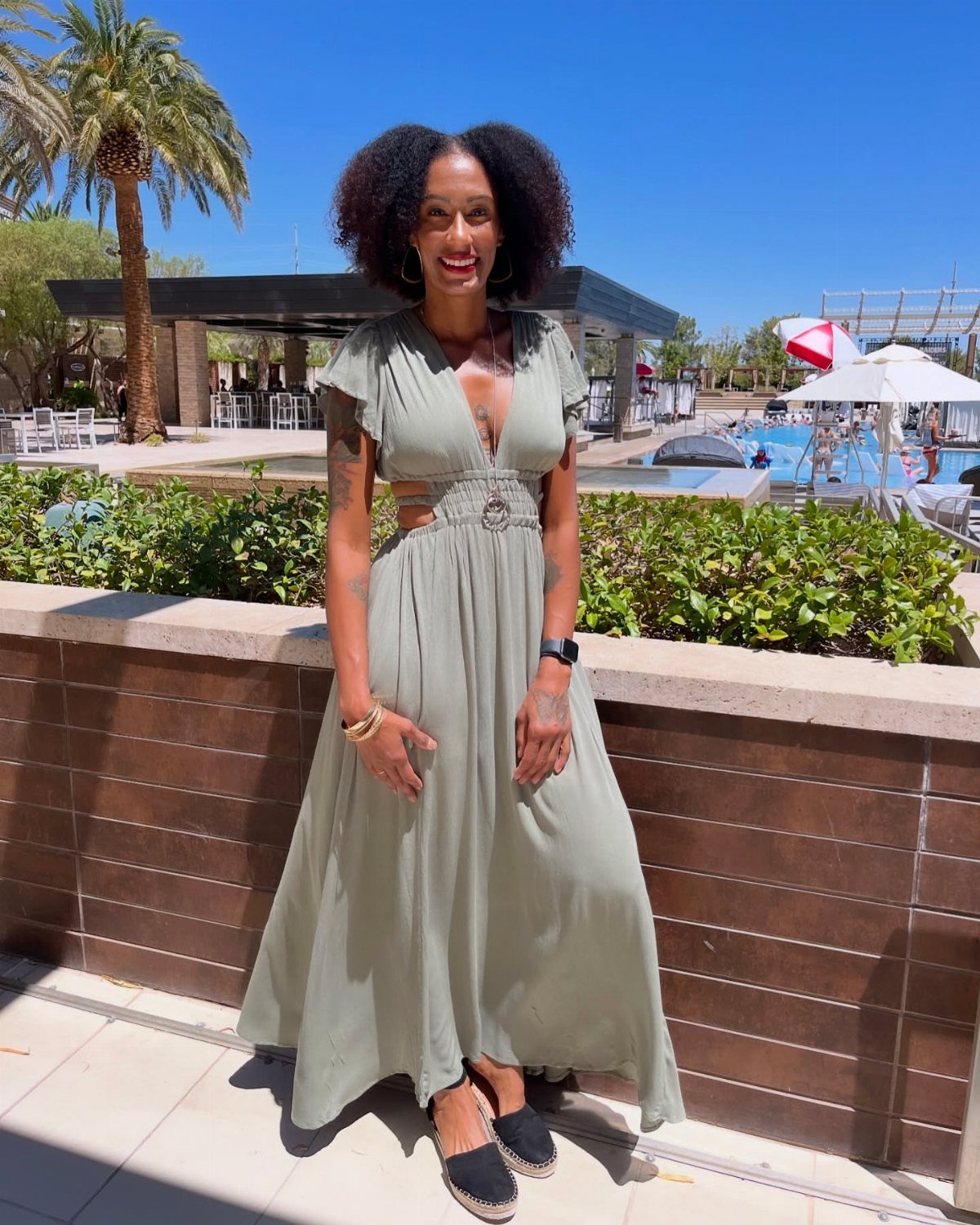 Woman in a sage green maxi dress standing at a sunny resort pool area