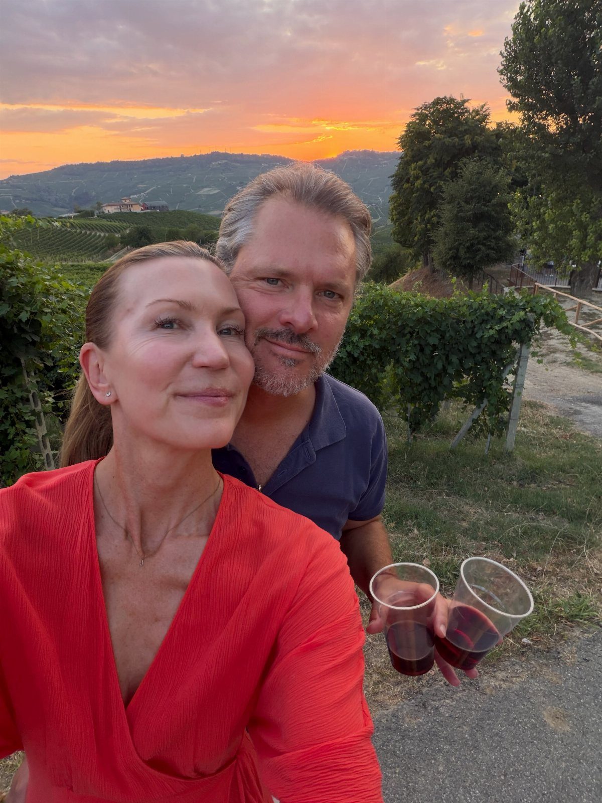Couple holding glasses of red wine in a vineyard during a sunset.