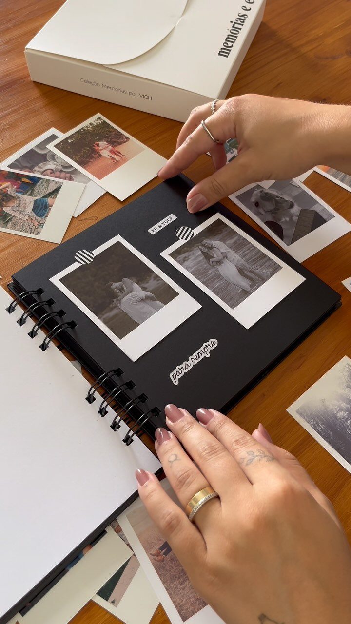 Hands arranging polaroid photos in a black spiral scrapbook on a wooden table