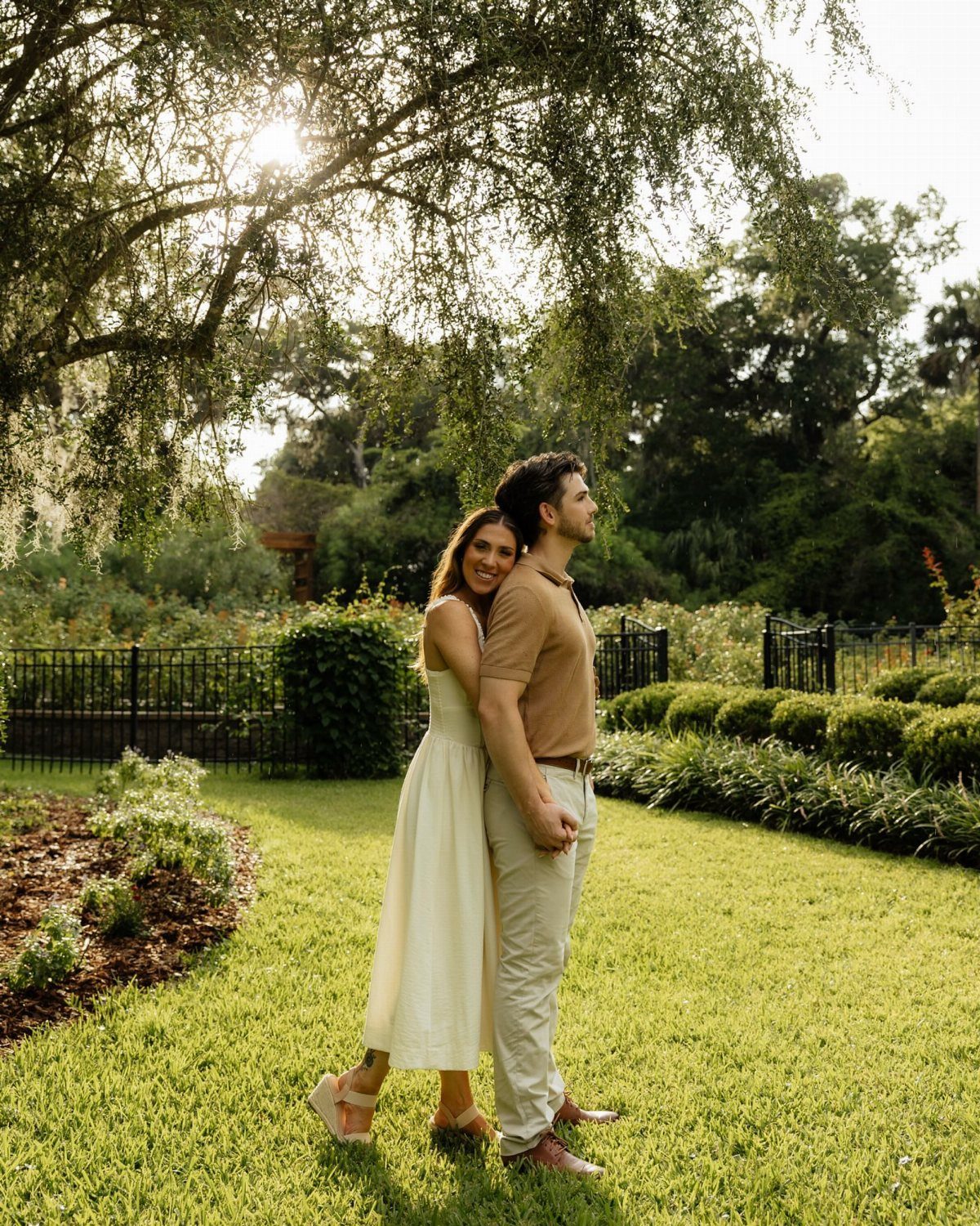 Couple embracing in a sunny botanical garden with manicured hedges and large trees.