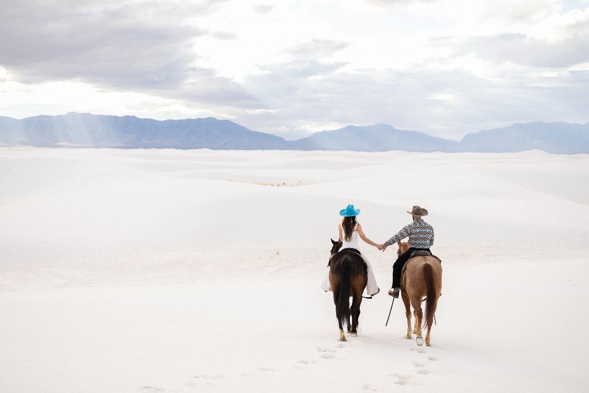 Couple riding horses and holding hands across a white sand desert landscape