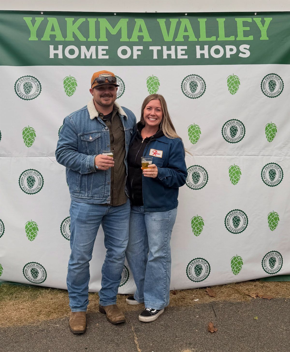 Smiling couple holding beer glasses in front of a Yakima Valley festival backdrop.