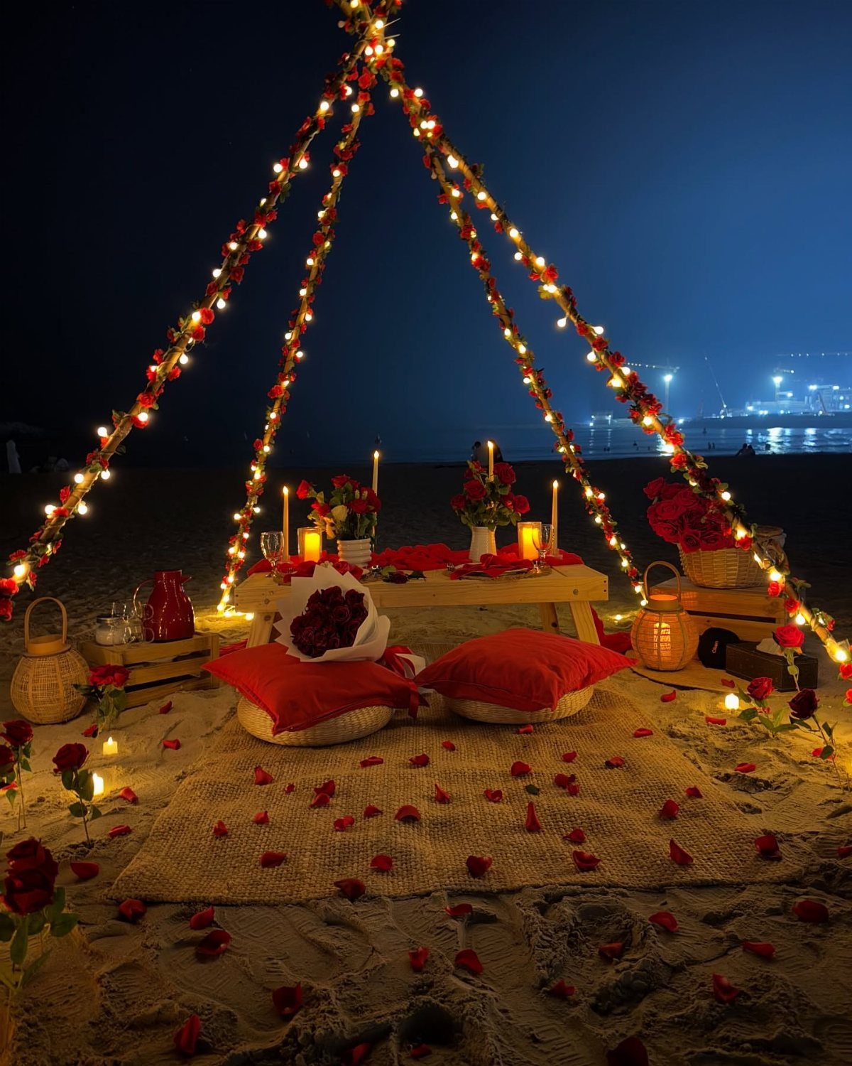 Romantic beach picnic setup with fairy lights, red cushions, and candles on sand.